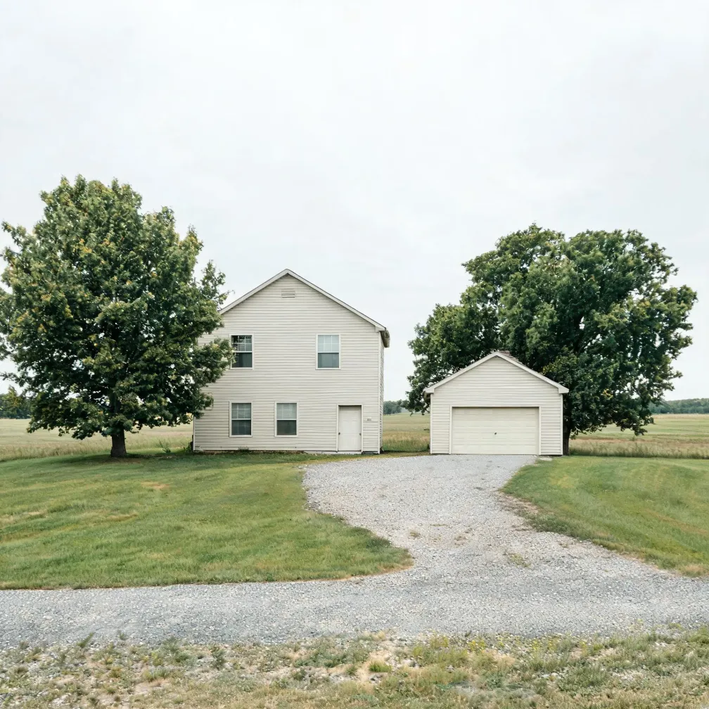 A two-story cream vinyl-sided house and a matching detached two-car garage stand on a flat grassy lot, flanked by two large deciduous trees, with a gravel driveway leading to the foreground under an overcast white sky.