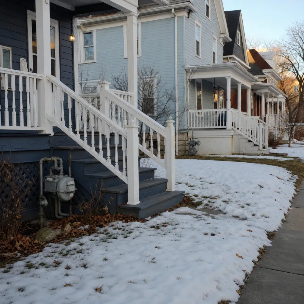 A row of wood-sided houses with white-railed front porch stairs, a gas meter beside the foundation, and patchy snow covering the lawn next to a concrete sidewalk.