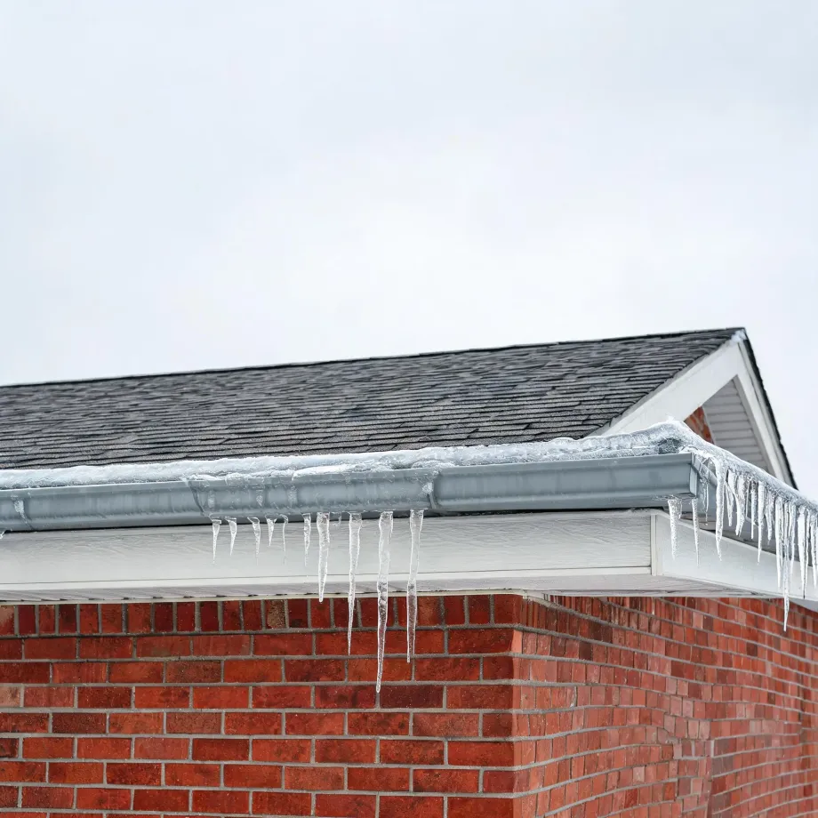 Corner of a brick house with asphalt shingle roof, white soffit and gray gutter, with icicles hanging from the gutter against an overcast sky.