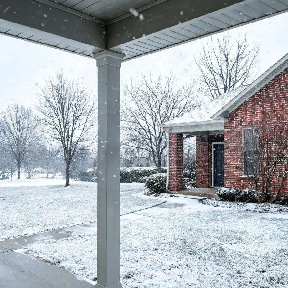 View from beneath a covered porch with a square white column toward a red brick house with a gabled entry, bare trees and shrubs, and snow falling on a snow-covered lawn and walkway.