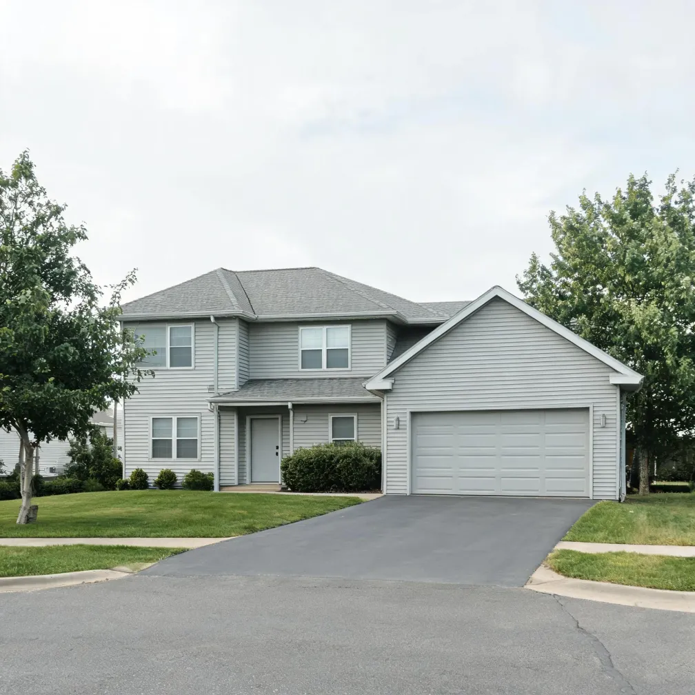 Two-story house with light gray horizontal siding, gray shingled roof, attached two-car garage, and a paved driveway leading to a street, with green lawn and deciduous trees on either side under an overcast sky.