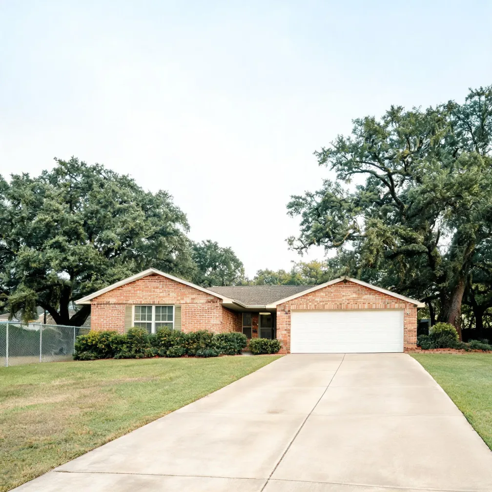 Single-story brick house with a two-car white garage door, shrubs along the front, a wide concrete driveway, mown lawn, chain-link fence at left, and large trees behind under an overcast sky.