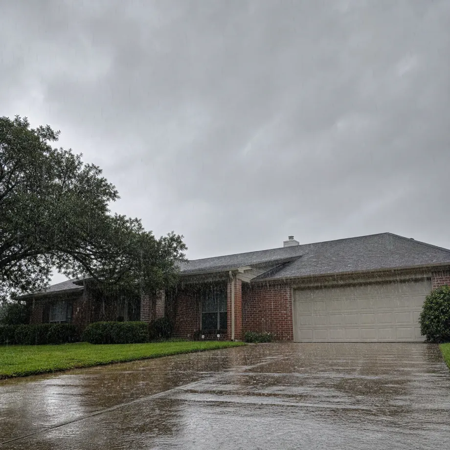 A single-story brick house with a two-car garage and shingled roof under a heavy overcast sky, with rain falling, water streaming from the eaves, and a wet driveway in the foreground beside a large tree and lawn.