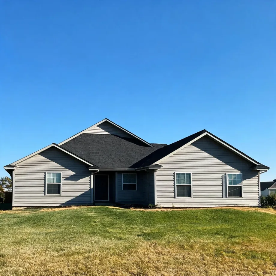 A single-story house with gray vinyl siding, dark shingled gable roofs, and white-trimmed windows, set behind a partly dry green lawn under a clear blue sky.