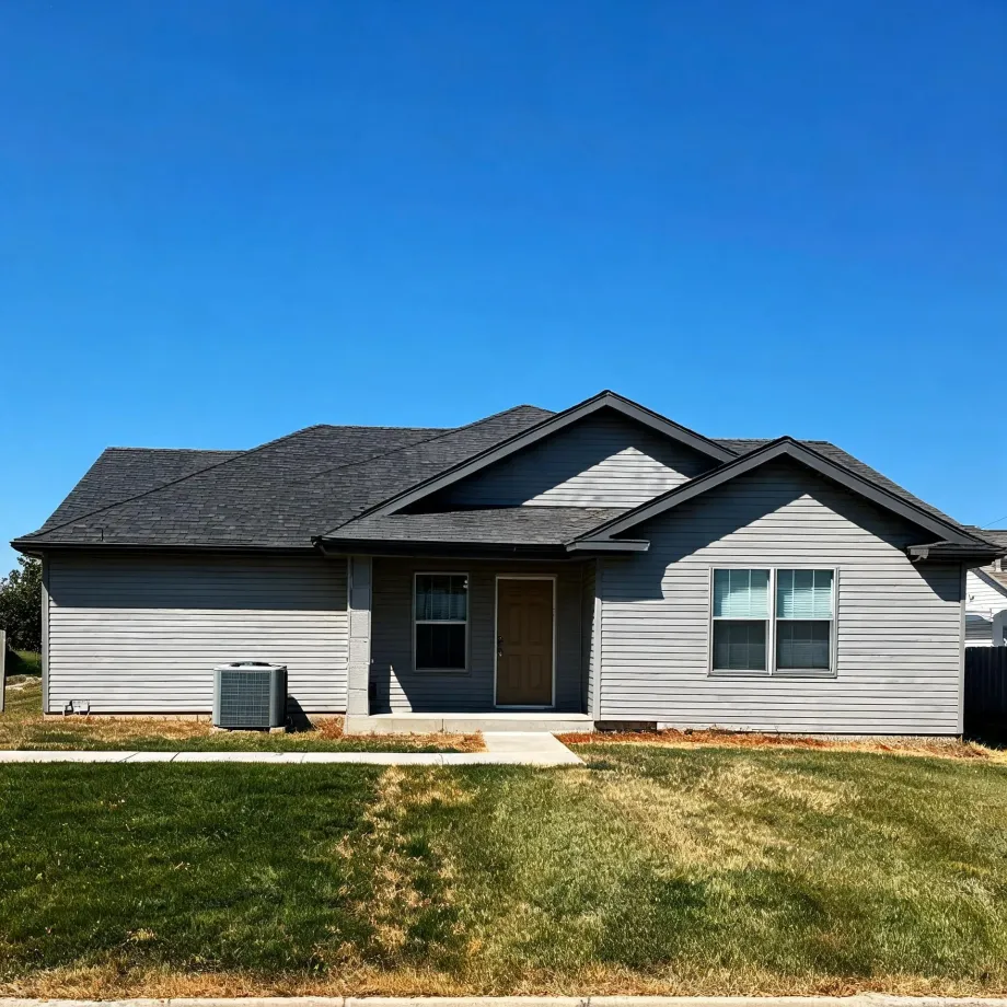 Single-story gray horizontal-siding house with dark shingled gables, a brown front door, front windows, an exterior air conditioning unit, and a patchy green and brown lawn under a clear blue sky.