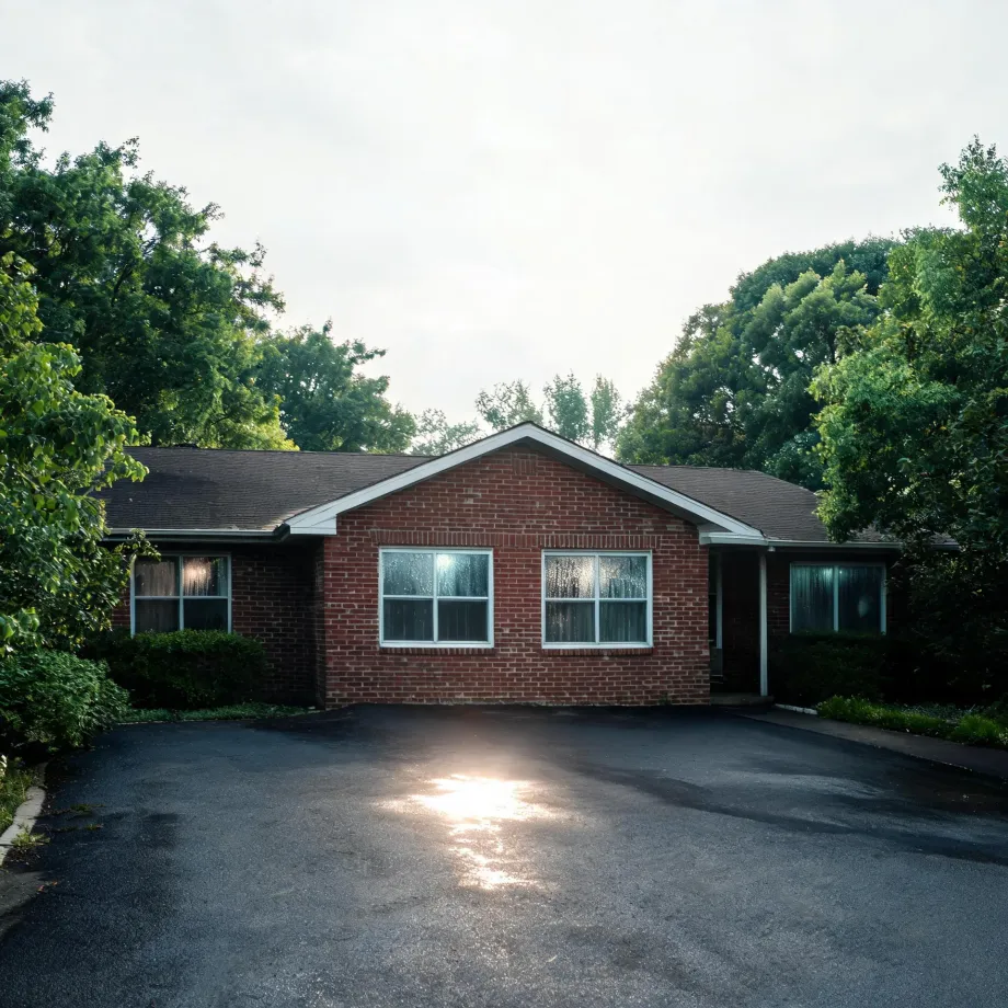 A single-story red brick ranch house with white-framed windows and a dark shingled roof, surrounded by leafy green trees, with a wide asphalt driveway in the foreground reflecting sunlight under an overcast sky.