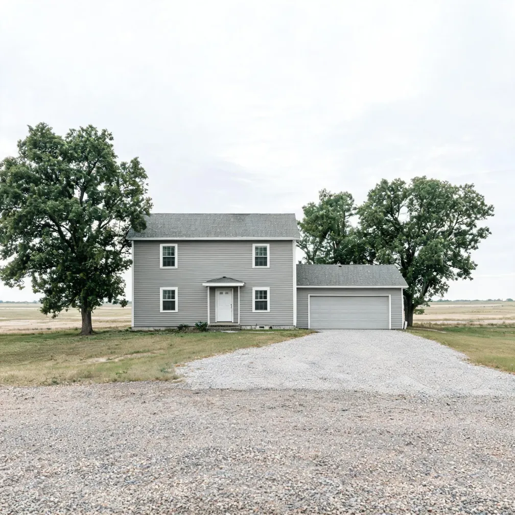 A two-story gray house with white-trimmed windows and an attached single-story garage, flanked by two large deciduous trees, with a wide gravel driveway in the foreground and flat open fields under an overcast sky.