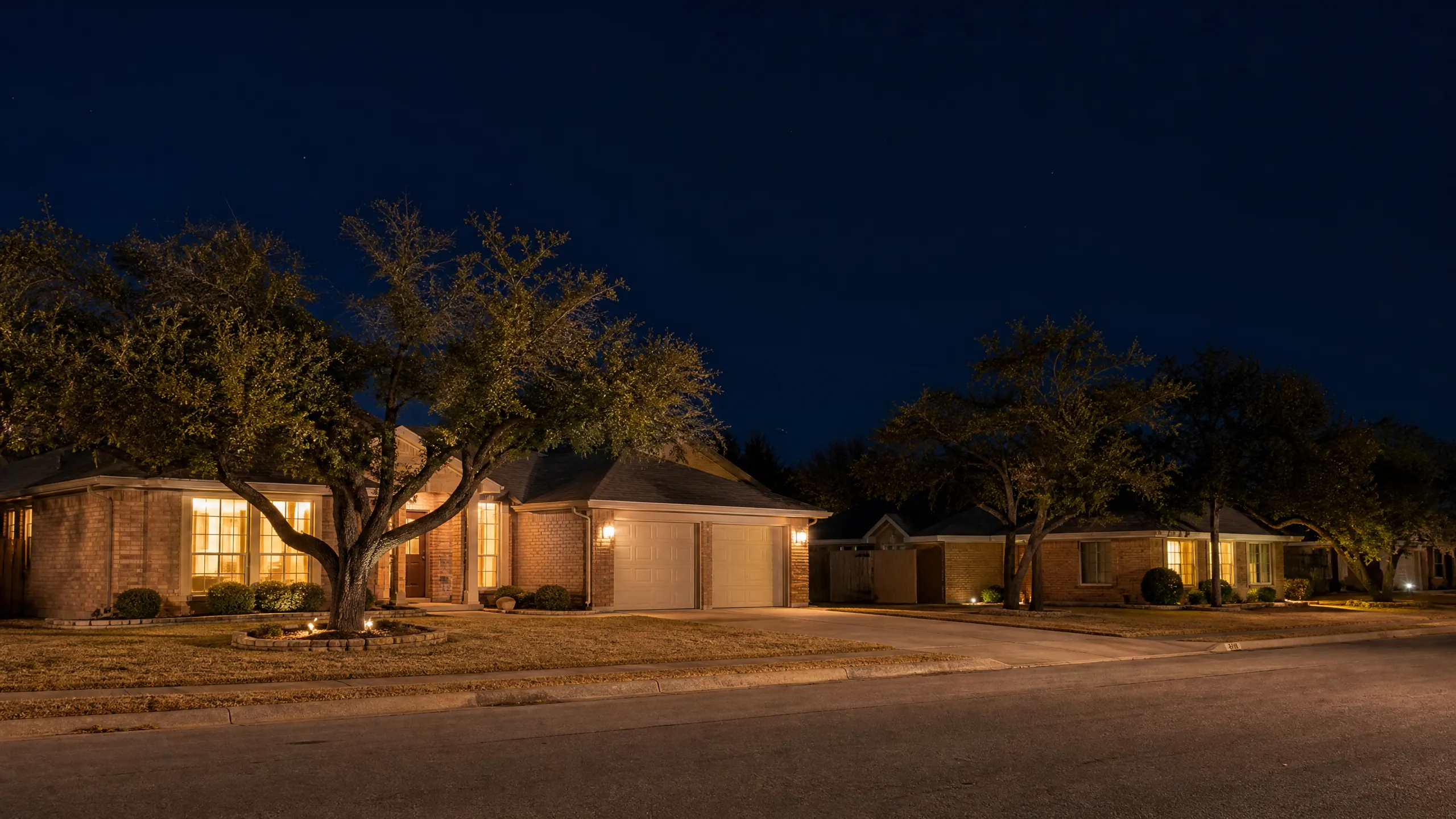 Two single-story brick suburban houses with attached garages and mature trees, photographed at night under a dark blue sky.