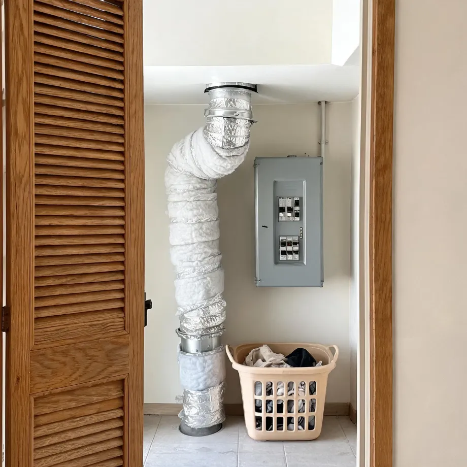 A closet interior seen through an open louvered wood door, with an insulated flexible duct running from the ceiling to the floor, a gray electrical breaker panel on the wall, and a tan laundry basket holding clothes on the tile floor.