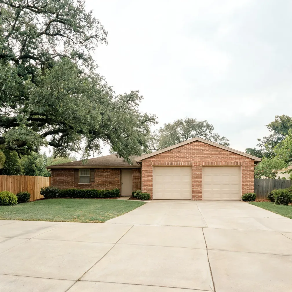 Single-story brick house with two separate white garage doors, a wide concrete driveway, mown lawn, wooden fences on both sides, and large oak trees under an overcast sky.