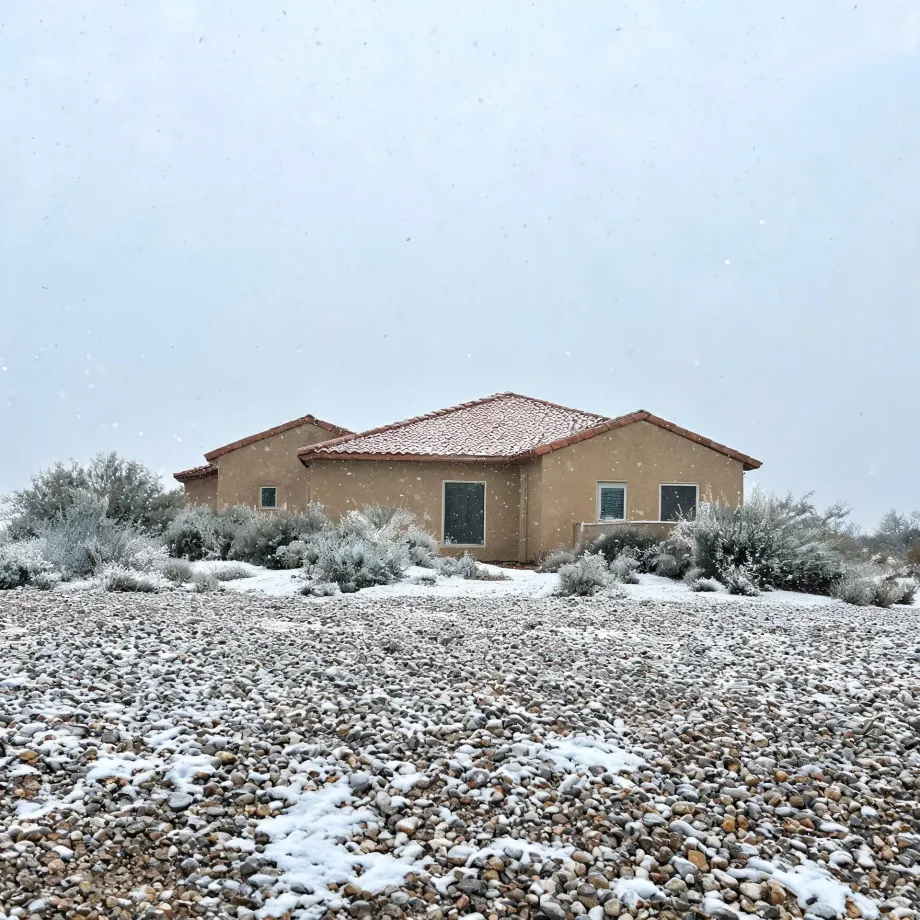 A single-story tan stucco house with a red tile roof behind snow-dusted desert shrubs and a gravel yard, under an overcast sky with falling snow.