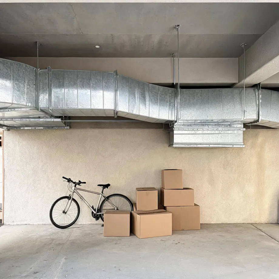 A bicycle leans against a beige stucco wall beside five stacked cardboard boxes in a concrete-floored space, with large galvanized metal air ducts suspended from the ceiling above.