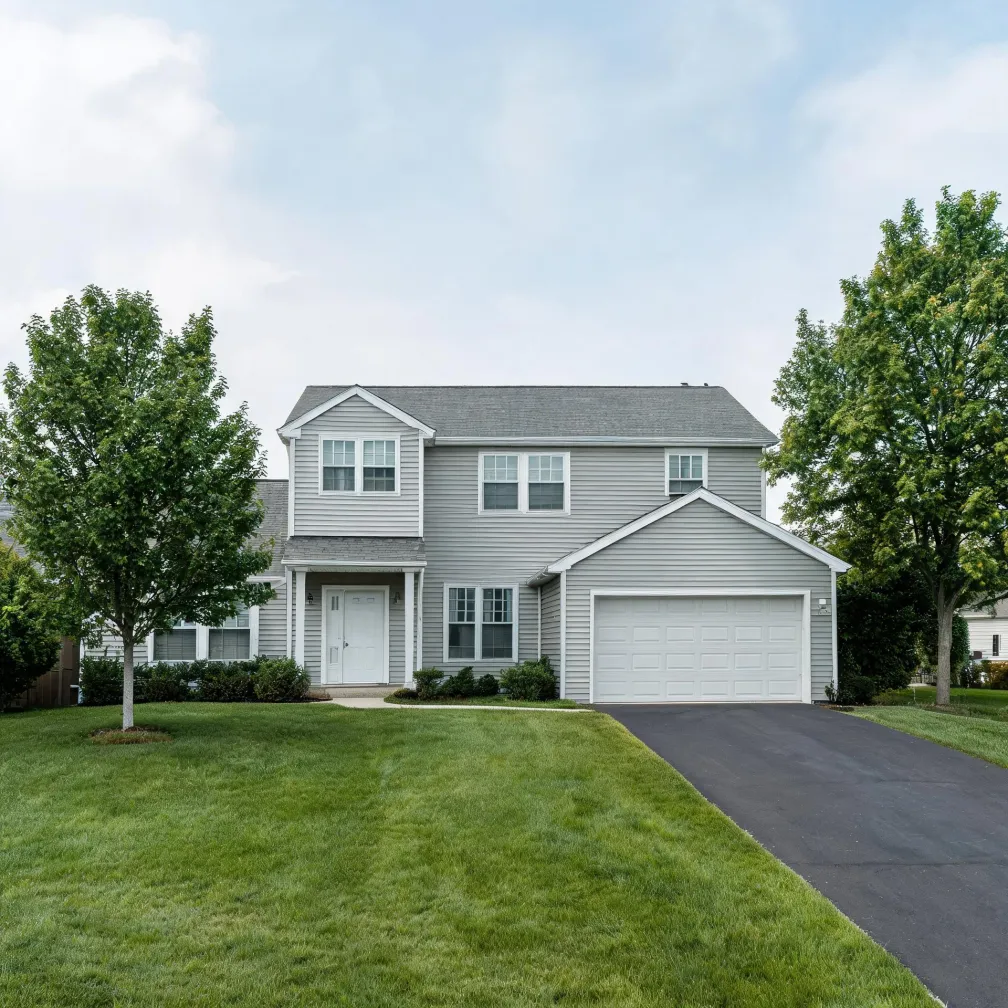 Two-story gray vinyl-sided house with white trim, a white front door, an attached two-car garage, an asphalt driveway, mown lawn, and deciduous trees on either side under a hazy sky.