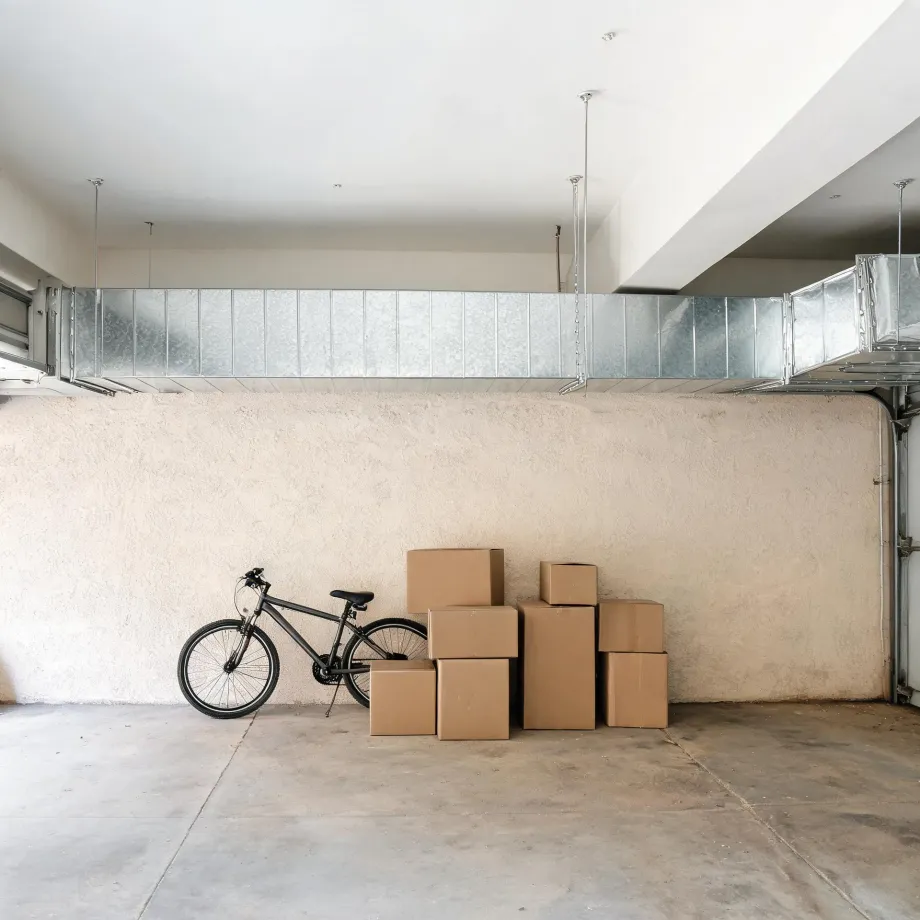 An empty garage interior with a textured beige wall, exposed metal HVAC ductwork near the ceiling, a black bicycle leaning against the wall, and a stack of cardboard boxes on the concrete floor.