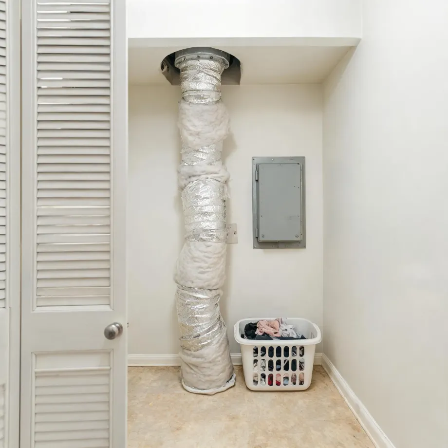 An interior alcove with an insulated flexible duct running from the ceiling to the floor, a gray electrical panel on the wall, a white laundry basket with clothes in the corner, and a louvered door at left.
