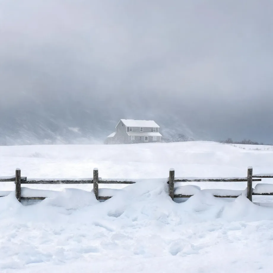 A two-story light-colored house stands on a snow-covered rise under a gray sky during blowing snow, with a partially buried wooden post-and-rail fence in the foreground.