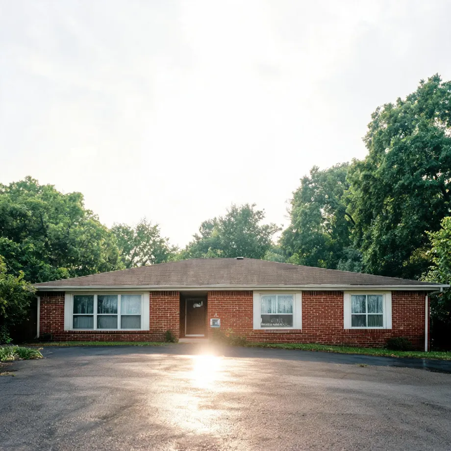 A single-story red brick ranch house with a low hipped shingle roof, white-trimmed windows and a central dark door, fronted by a large asphalt parking area with sun glare, surrounded by leafy green trees under a bright hazy sky.