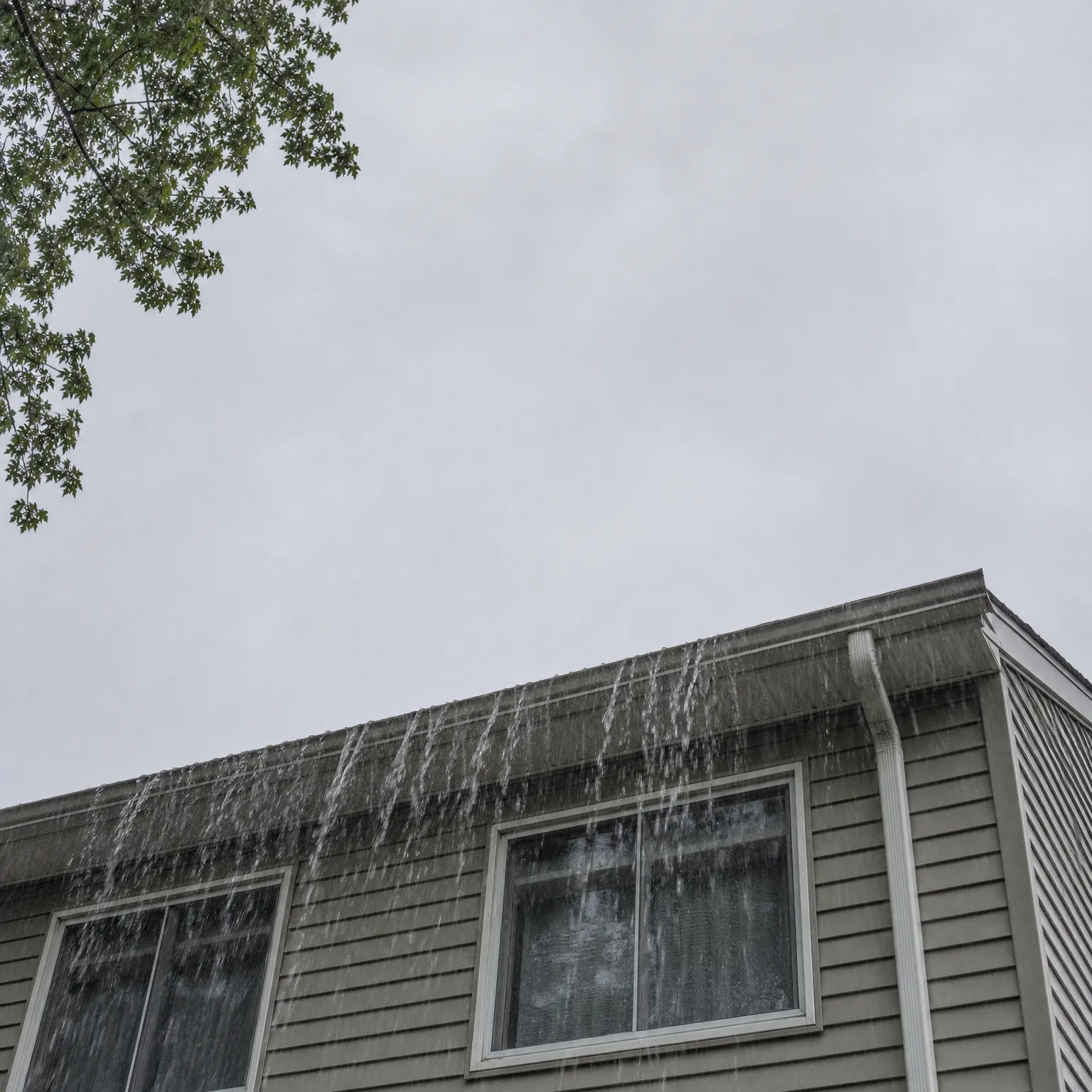 Upward view of the corner of a two-story house with horizontal lap siding and white-framed windows, with water spilling over the roof edge and gutter against an overcast sky, and tree branches in the upper left.