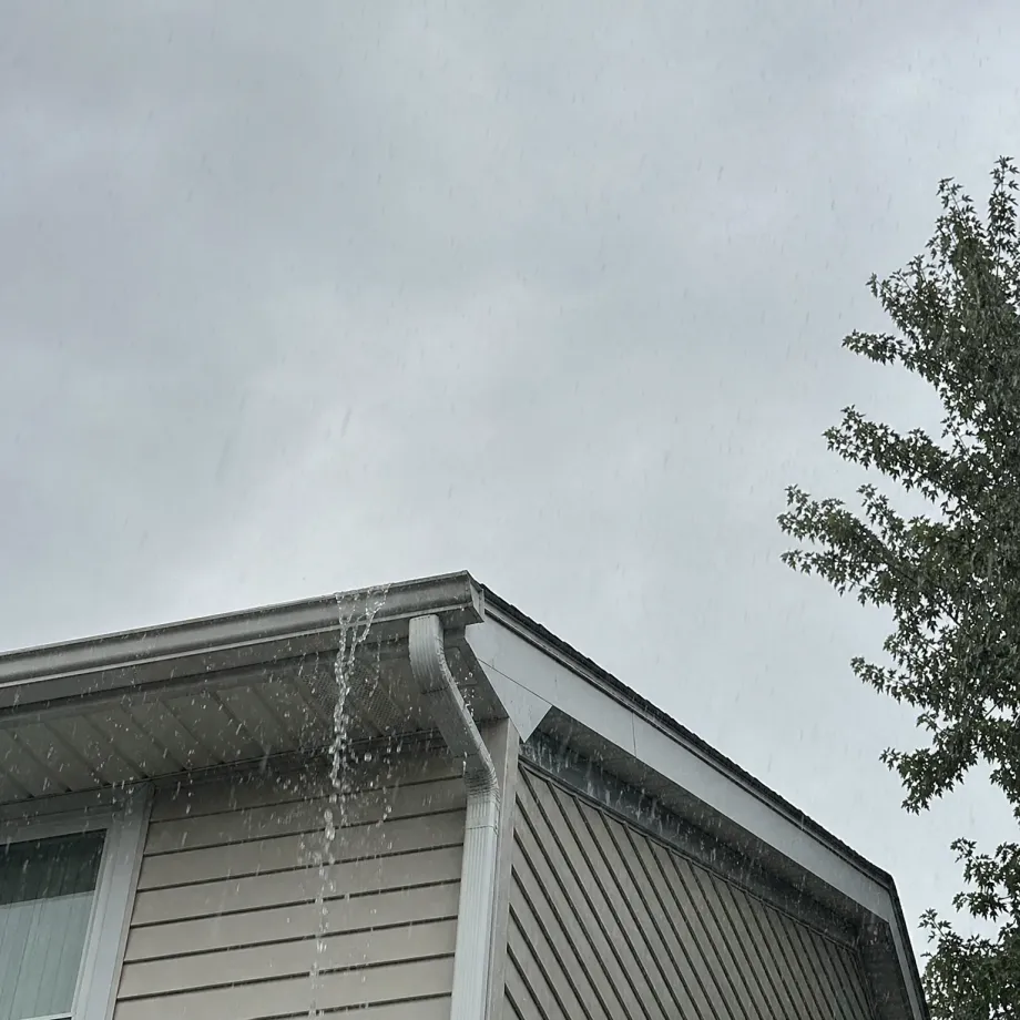 Low-angle view of the corner of a house with vinyl siding and a gutter overflowing water during rain, under an overcast gray sky with a leafy tree at right.