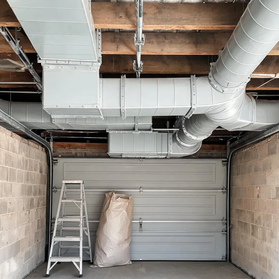 Interior of a garage with concrete block walls and a closed sectional door, showing large sheet-metal rectangular and round ductwork running along the exposed wood joist ceiling, with an aluminum step ladder and a large paper bag on the floor.