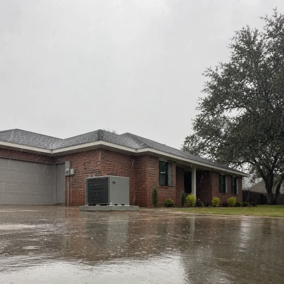 A single-story red brick house with a gray shingled roof, attached two-car garage, and exterior HVAC units on a concrete pad, seen from a wet driveway during rain with a large oak tree at right.
