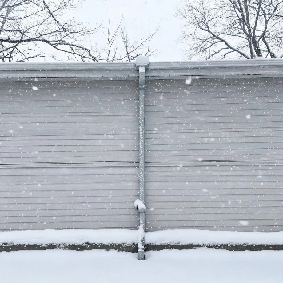A gray horizontal-lap sided wall with a vertical downspout, photographed during falling snow, with snow-covered ground and bare tree branches above the roofline.