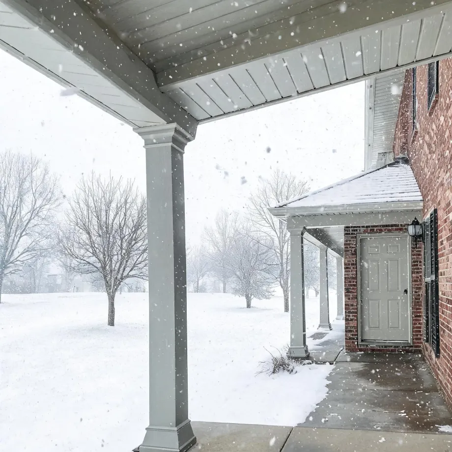 View from under a covered porch with white columns toward a snow-covered lawn and bare trees, with a red brick house wall and light-colored entry door at right during falling snow.