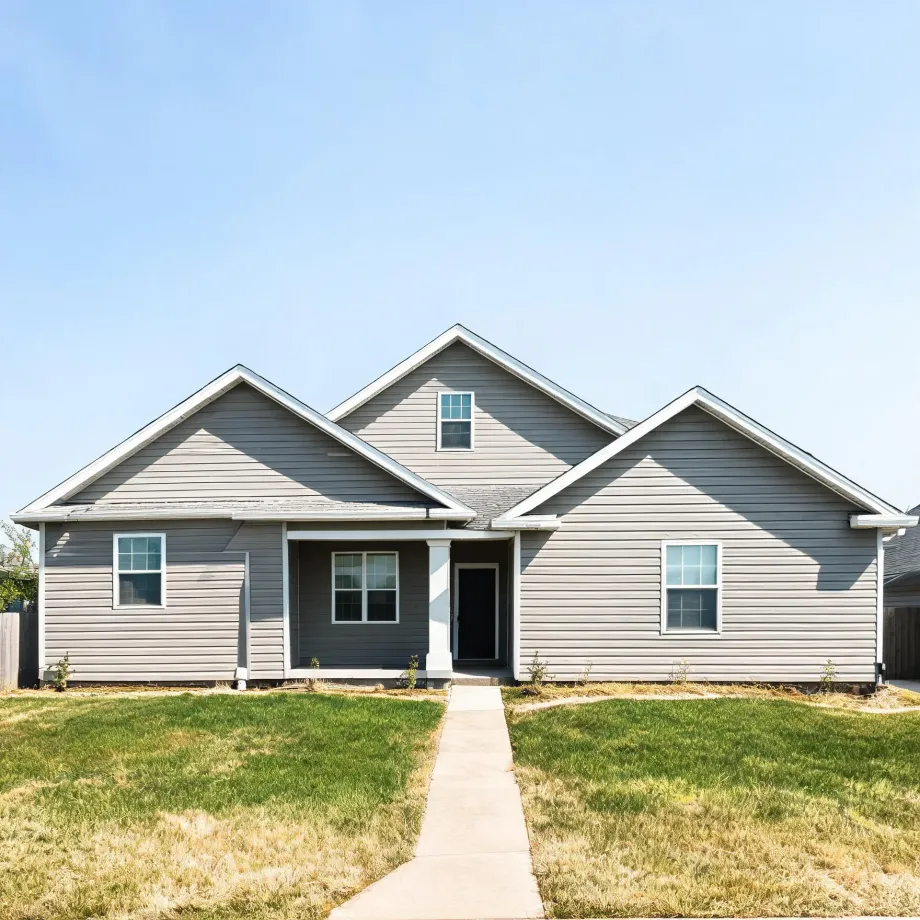 A single-story gray horizontal-siding house with multiple front-facing gables, white-trimmed windows, a dark front door, and a concrete walkway crossing a partly dry lawn under a clear blue sky.