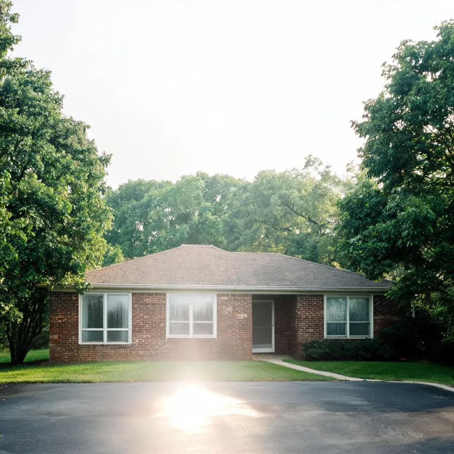 A single-story red brick house with a hipped shingle roof, large windows and a recessed front door, set behind a lawn and asphalt driveway with sun flare, framed by leafy green trees.