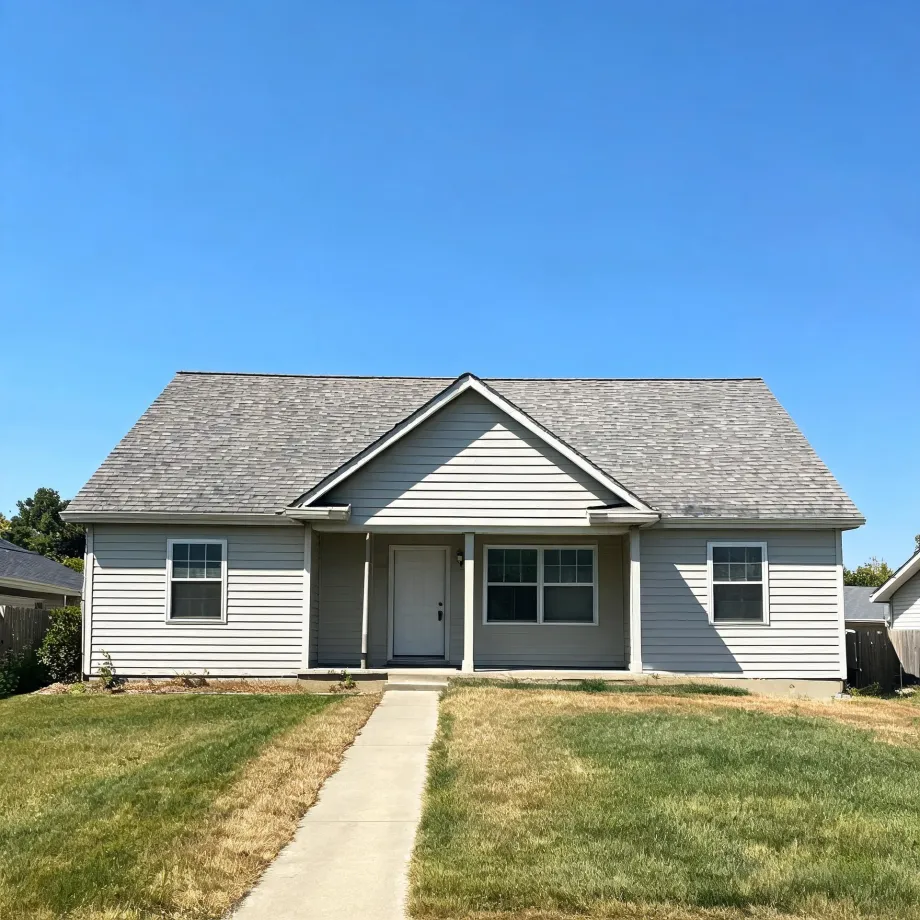 Single-story house with light gray horizontal siding, gray shingled roof, small front porch with square columns, and a concrete walkway crossing a partly dry lawn under a clear blue sky.