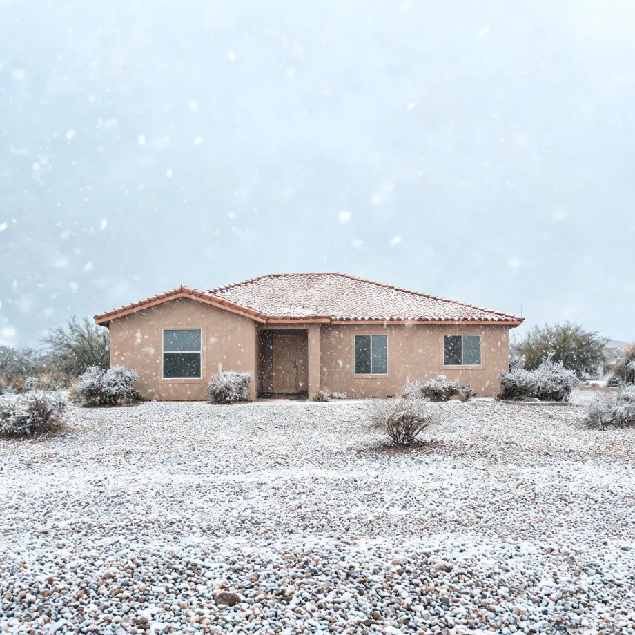 A single-story tan stucco house with a tile roof during falling snow, surrounded by low desert shrubs and a gravel yard dusted with snow.