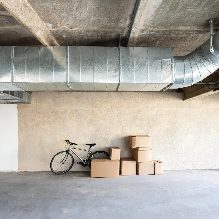 Interior of a concrete-floored garage or basement with large galvanized metal ventilation ducts under the ceiling, a bicycle leaning against a beige stucco wall, and several stacked cardboard boxes beside it.