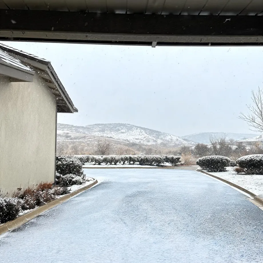 View from under a covered walkway of a snow-dusted driveway lined with trimmed shrubs, a stucco building with a tiled roof at left, and low hills under an overcast sky.