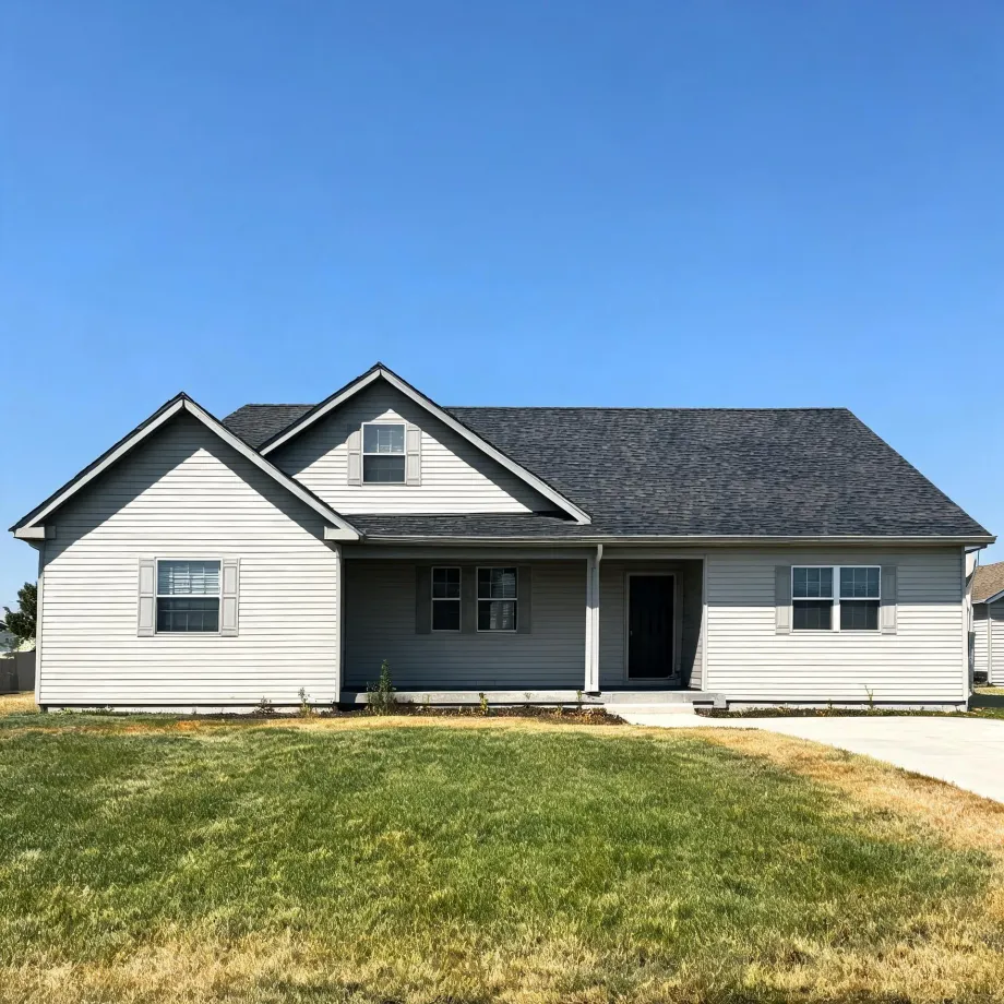 A single-story house with light horizontal siding, gray shingled roof, gabled dormer, shuttered windows, and a small covered entry porch, fronted by a patchy green lawn and a concrete driveway under a clear blue sky.