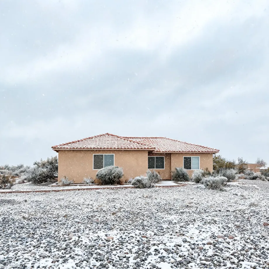 A single-story tan stucco house with a red tile roof stands behind a snow-dusted gravel yard and low desert shrubs under an overcast sky with falling snow.