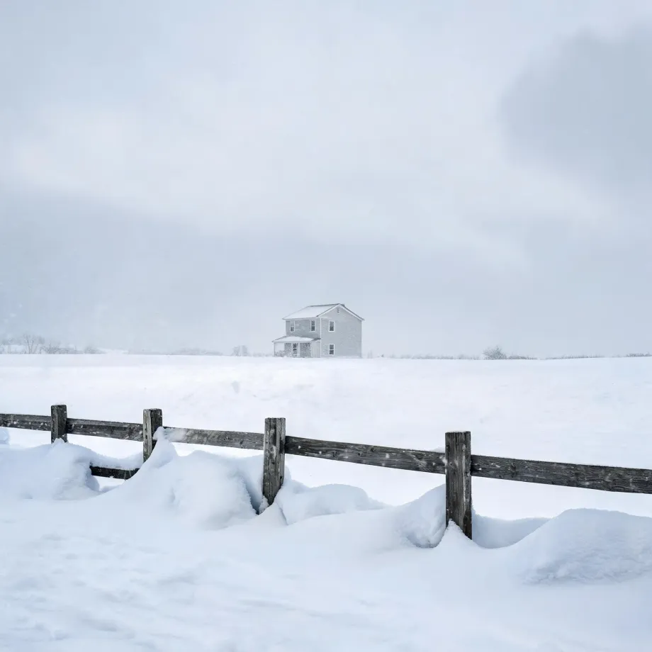 A two-story light-colored house stands alone in a snow-covered field under a pale overcast sky, with a weathered wooden post-and-rail fence partly buried in snowdrifts in the foreground.