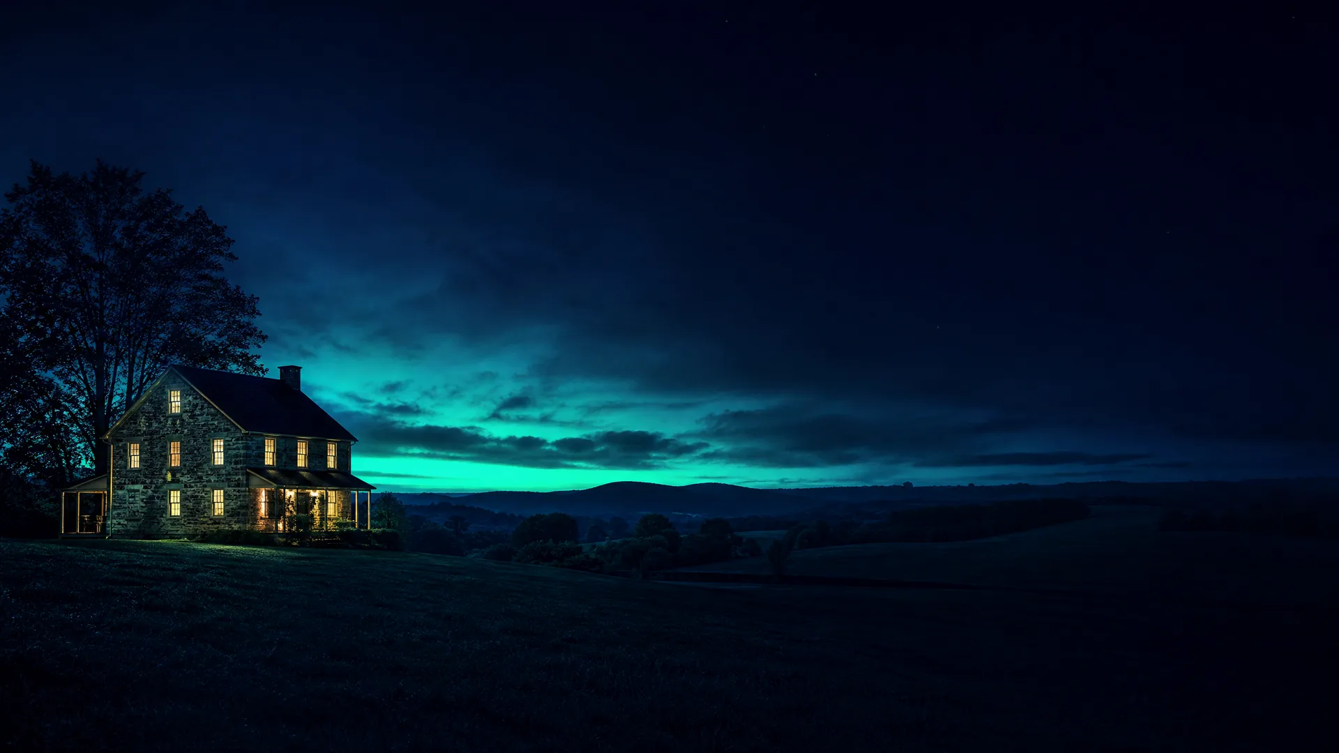 A two-story stone farmhouse with lit windows stands on a dark grassy hilltop beside tall trees, under a twilight sky with a band of teal glow above distant low hills.