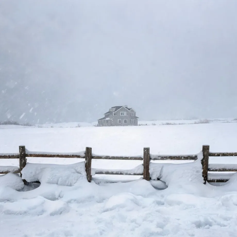 A two-story gray house stands alone in a snow-covered field during falling snow, with a snow-laden wooden post-and-rail fence in the foreground and a pale, hazy sky above.