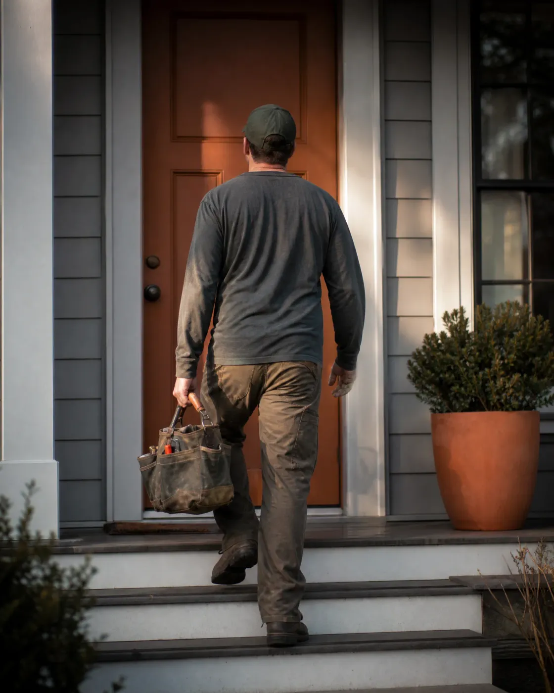 A man in a cap, long-sleeved shirt and work pants carries a canvas tool bag up concrete steps toward a wooden front door of a gray-sided house, with a shrub in a terracotta pot to the right.