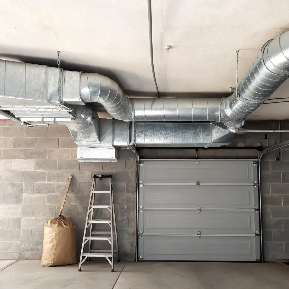 Galvanized rectangular and round HVAC ductwork suspended from a ceiling in a concrete block garage, above a gray sectional garage door, with a step ladder and a paper bag against the wall.