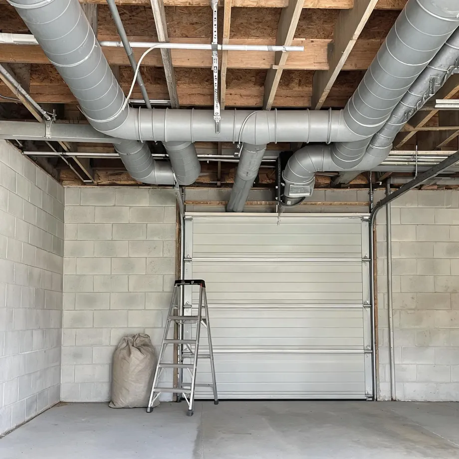 Interior of a concrete block garage with a closed sectional door, exposed wood joists overhead carrying large round metal ducts and pipes, a folding aluminum step ladder and a tied cloth sack on the bare concrete floor.