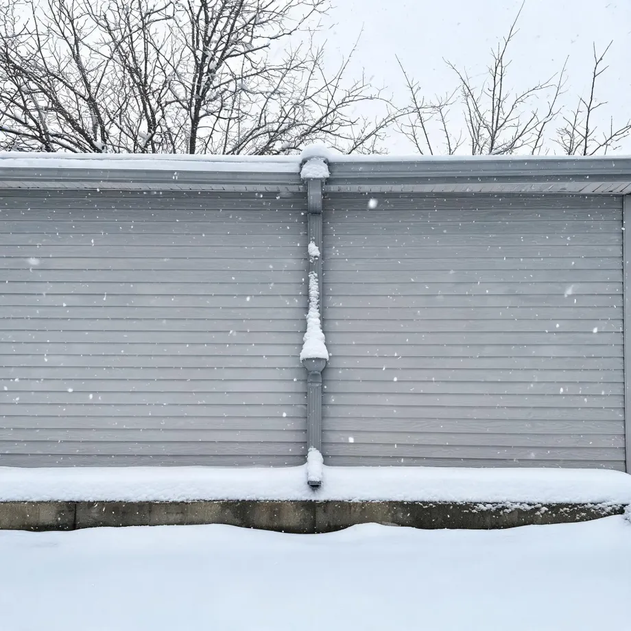 Gray horizontal vinyl siding on a building wall with a central downspout coated in snow, a snow-covered concrete foundation ledge and ground below, and bare tree branches against a pale sky above while snow falls.