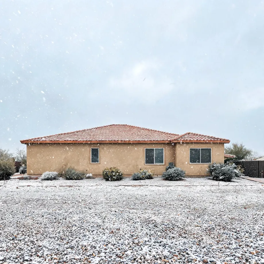 A single-story tan stucco house with a red tile roof during falling snow, with light snow covering a gravel yard and low desert shrubs and cacti in front.
