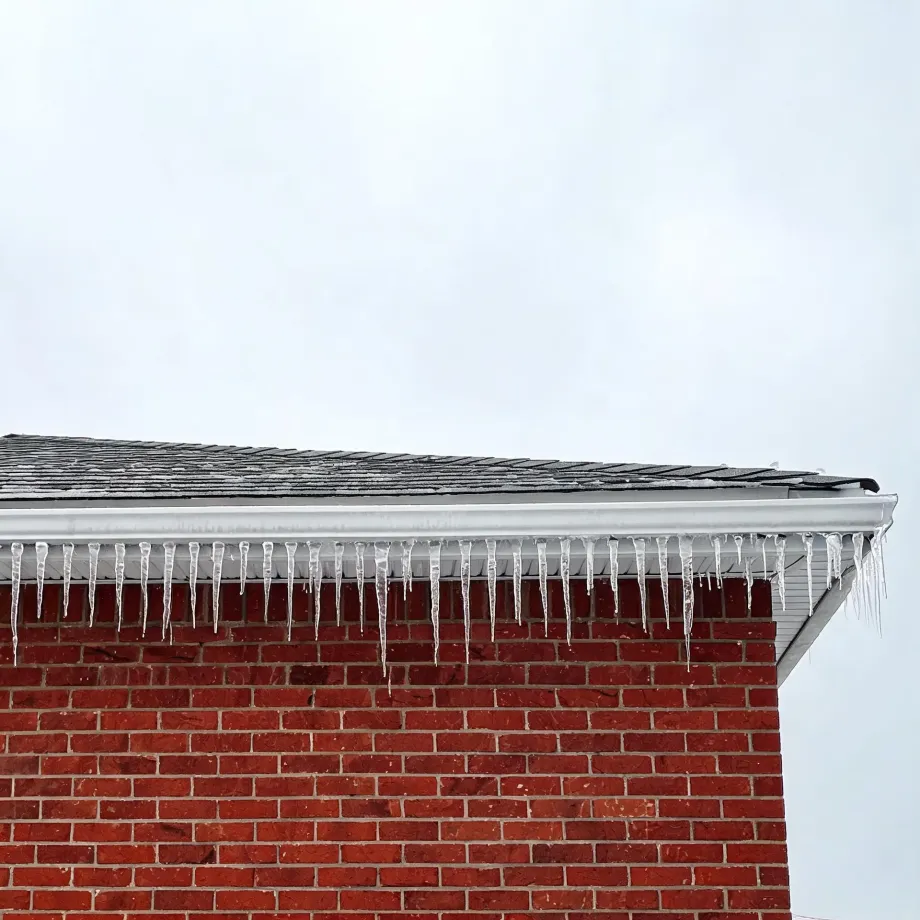 Icicles hang from the white gutter along the edge of a shingled roof on a red brick building under an overcast sky.