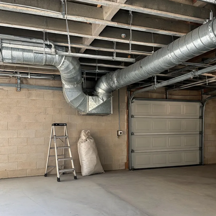 Interior of an empty garage with concrete block walls, a closed sectional garage door, exposed wood joists with large galvanized round and rectangular ductwork, and a step ladder and filled sack against the wall.