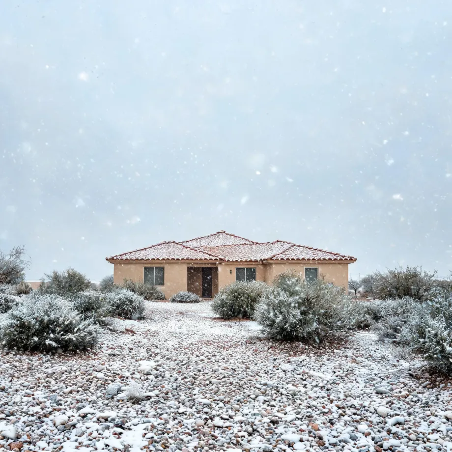 A single-story tan stucco house with a red tile roof stands behind snow-dusted desert shrubs and a gravel yard during falling snow under a pale overcast sky.