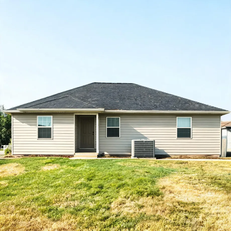 Single-story house with beige horizontal siding, dark shingled hip roof, a brown front door, three windows, and an air conditioning unit, fronted by a patchy green and dry lawn under a clear sky.