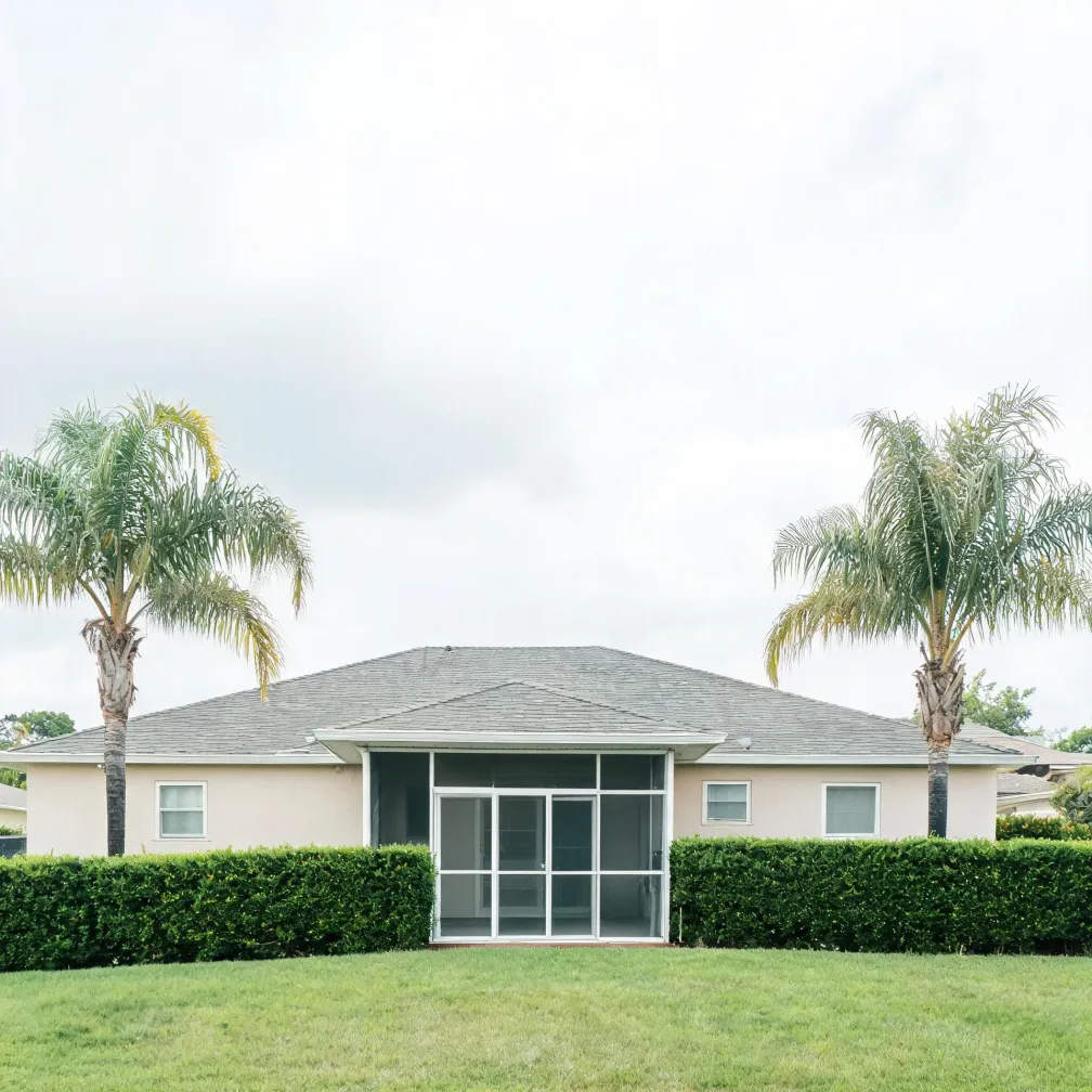 Rear exterior of a single-story stucco house with a gray shingled hip roof and a screened porch, flanked by two palm trees, behind a trimmed hedge and a mown lawn under an overcast sky.