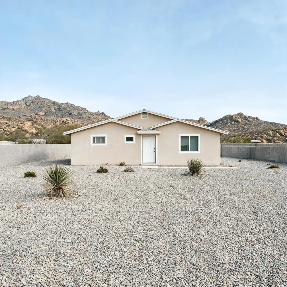 A single-story beige stucco house with a gabled roof, white front door and white-framed windows, set behind a gravel yard with scattered yucca plants, a low concrete block wall, and rocky hills in the background.