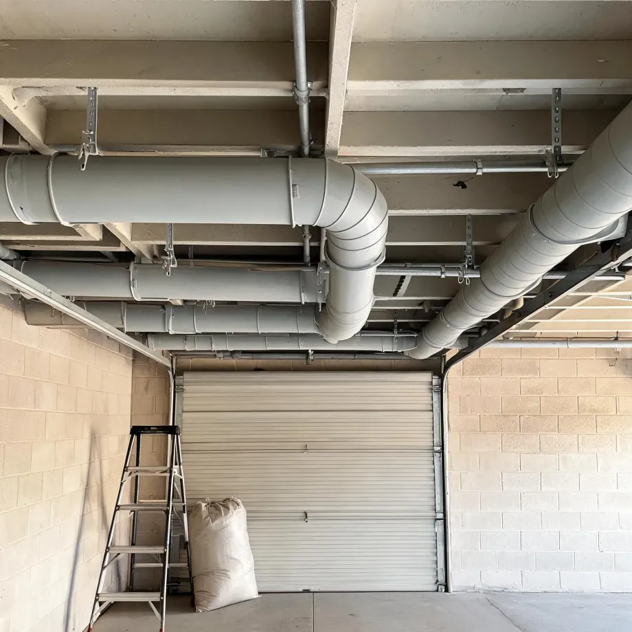 Interior of a garage with concrete block walls, a closed roll-up door, insulated round ductwork and conduit suspended from the concrete ceiling, a step ladder and a filled sack on the floor.